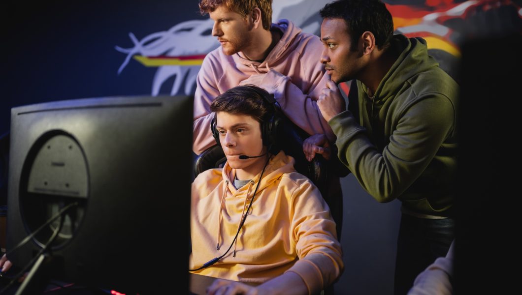 Interracial team standing near gamer playing video game on computer in cyber club